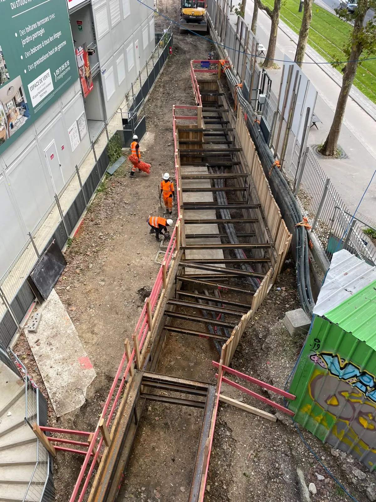 Tranchée blindée le long d’un bâtiment, accès par escalier de chantier.
